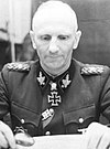 A black-and-white photograph of a man stiing at a desk, wearing a military uniform and neck order, in shape of an Iron Cross.