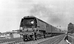A posed side-and-front view of a large 4-6-2 steam locomotive with a tender. The locomotive boiler is hidden by a casing of flat metal side sheets.