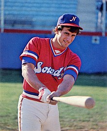 A man in a red baseball jersey and blue cap posed holding his bat forward