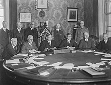 Six white men sitting around a conference table, with five more standing behind them. One of the seated men, third from the left, is sitting in a much more ornate chair than the others; he is the Prime Minister of Canada, William Lyon MacKenzie King.