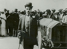 Frontale Schwarzweißfotografie eines jungen Mannes mit einer Menschenmenge im Hintergrund. Der Mann trägt einen Melonenhut und einen kurzen Schnurrbart. Er hat eine Ansteckblume an seinem Anzug und hält links einen Gehstock. Rechts steht ein Oldtimer mit offenem Fahrersitz.