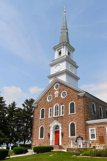 The front of a brick chapel with a white steeple, taken on a sunny day with a cloudy blue sky.