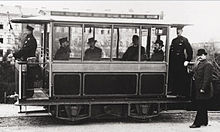 Ein 5 Meter langer Straßenbahnwagen mit offenem Führerstand, besetzt mit Fahrer, Schaffner und vier Passagieren