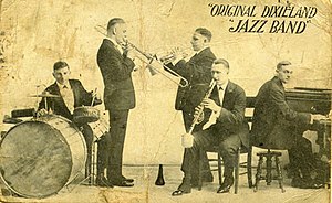 A jazz band playing: A drummer on the left behind a drum set, a trombonist next to him facing right. A cornetist standing behind the trombonist facing left, and a clarinetist sitting on a chair in the front. A pianist sitting on the far left, facing right.