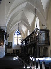 Frontale Farbfotografie aus dem Innenraum einer Kirche mit dunklen Bänken und dunkler Empore an der rechten Seite. Links hängen an den Wänden und Pfeilern Gemälde. An der hinteren Wand ist ein farbiges Kirchenfenster und von dem weißen Gewölbe hängen Kronleuchter.