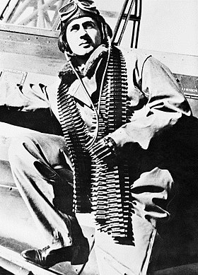 Three-quarter portrait of moustachioed man in flying suit with a belt of machine-gun ammunition slung over his shoulders, leaning against an aeroplane