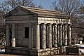 Light colored stone mausoleum flanked by four columns. Door is aged metal.