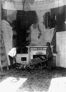 a black and white image of a room ravaged by fire with a man holding a wheelbarrow full of debris in front of a fireplace