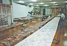 A well-lit workshop. A length of white cloth patterned with bingata is stretched along the length of the room, and a person works on it in the background.