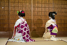 A maiko (on the left) and a geisha (on the right) facing away from the camera, sat on a tatami mat.