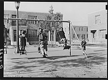 A group of approximately four Black children play on a swing set surrounded by buildings in 1938..