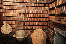 A photograph of two walls containing bookshelves filled with antique volumes; in the foreground a set of globes and a map screen