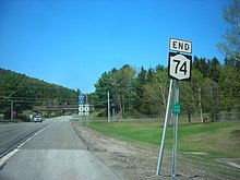 Ground-level view of a road and an associated road sign. More roadsigns and an overhead bridge are visible in the distance.