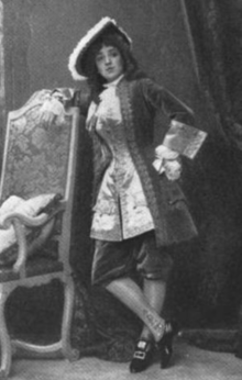 A white woman in a corseted costume, resembling the dress of an 18th-century European man, with a brimmed hat, a long coat, an embroidered waistcoat, knee-length reeches, and silk stockings; she is standing next to a chair, with one hand on her hip