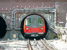 Front view of a small profile train emerging from the tunnel north of Hendon Central on the Northern line, showing the small gap between the train's curved roof and tunnel's "tube".
