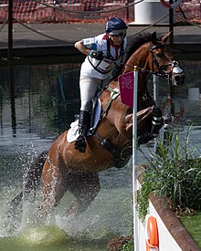 white-attired rider on a chestnut horse which is leaping out of a water jump