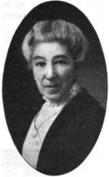 An older white woman, grey hair in a bouffant updo, wearing a high-collared white lacy blouse and a dark jacket.
