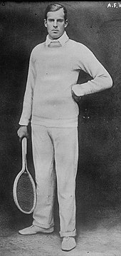 A man in white, with long pants and a sweater, holding a wooden racket in his right hand, looks into the camera
