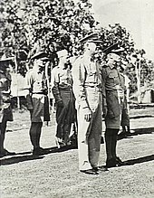 Full-length, three-quarter-angle, outdoor portrait of half a dozen or so men in light tropical military uniforms with headgear, standing to attention. Two of the men are in the foreground and the rest in a row behind them.