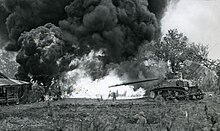 tank shooting flame into foliage and pillbox, creating a plume of smoke.