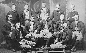 Baseball players are posing for a photograph, five men standing, five men sitting on chairs, and two are sitting on the floor.