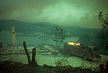 A fire truck burns in the driveway of a destroyed home with a view of a lake in the background; the sky is green-gray with smoke