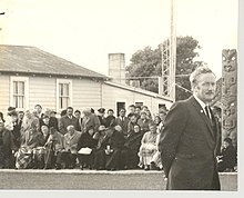 Duncan MacIntyre standing in the foreground at Raukawa Marae in Ōtaki 1971 at a gathering with rows of people sitting in the background and a carved post to one side.