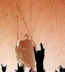 Image of the band's vocalist using stilts and wearing a long robe made of black feathers. In his hands are two elongated stilts, which he is holding aloft. The background of the stage is illuminated with orange stage lighting, and in the foreground numerous fans can be seen giving the 'devil-horns' signal.