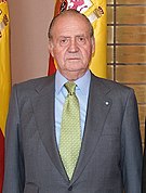 King Carlos in a grey jacket, light blue shirt, yellow tie, standing indoors in front of the Spanish flag
