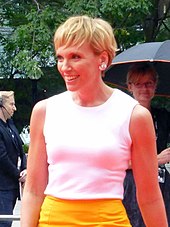 The 42-year-old is shown in a half-body shot. She wears a white top above a yellow skirt. She is smiling and facing to her left with her hair short, fair to light brown. Behind her to our left is a man wearing headphones and a microphone, while at the right is a woman holding an umbrella.