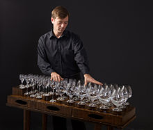 A man, Robert Tiso, has his hands on the rim of several wine glasses. Sound is produced by running a wet finger over the rim of wine glasses filled to different levels with water.