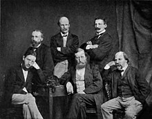 A group of six middle-aged men in dark jackets and ties, three seated and three standing