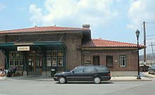 Entrance to a train station, signs read "Hastings-on-Hudson", "Station Cafe", "MTA Metro-North Railroad". Two people are sitting out front drinking coffee, and a black Mercedes wagon is parked.