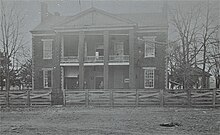 A sepia-tint black and white photograph of the front of an old two story brick building with a large porch and portico behind a wooden fence. A man stands in the doorway, a small painted sign advertising a lawyer's services is hung near the front door, and a gazebo is located to the right of the building.