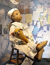 Doll in sailor suit seated on small chair; walls behind covered in pinned-up letters