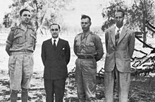 Four men pose awkwardly for a photograph. Two wear shirt-sleeve uniforms and the other two wear suits. All are bare-headed.