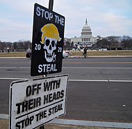 Two signs posing as street signs beside a road. In the background, the U.S. Capitol building is visible. The top sign is black with the top text saying "STOP THE", and the bottom text saying "STEAL" in all-caps; "stop the steal" was a common chant after the 2020 election. In the center, a skull with Donald Trump's hair is between the numbers '20' and '20', meaning the 2020 presidential election. The bottom sign reads, in all-caps, "OFF WITH THEIR HEADS. STOP THE STEAL".