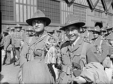 Two Maori men wearing military uniforms smile at the camera, surrounded by other soldiers in front of a building