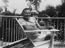Photograph of a man reclining on a chair on a porch, smiling at the camera and holding a drink.