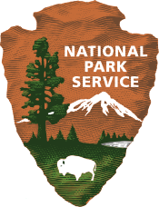 The National Park Service logo depicts an outline of a brown, jagged arrowhead, embedded in which are the silhouettes of a large green tree, a white mountaintop, and a grazing white bison. The words "National Park Service" hover above the mountaintop.