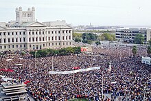 A crowd of thousands stands in a plaza. Many carry large banners.