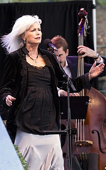 Singer Emmylou Harris with her arms extended, and singing into a microphone.