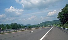 An expressway, photographed from its right shoulder, continuing north toward a green hillside in the center of the image, where it curves leftward. There is another hillside off to the left in the distance.