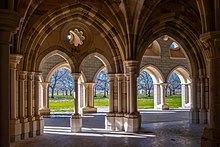Multiple interior and exterior stone arches frame a view looking out from a chapter house, revealing a view of orchard trees beyond an expanse of bare ground.