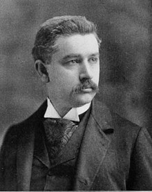 A man with dark hair and a mustache wearing a dark jacket and vest, patterned tie, and white shirt