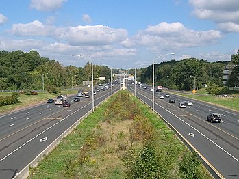 Both sides of a highway with a grass plot in the middle of the roads. Street lamps surround the middle, and several cars are on the roads. The roads have an HOV diamond on them.