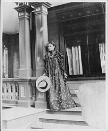 Likelike's daughter, standing on a porch in a long dress and holding a hat