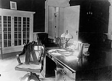 A black and white photo of desk and chair. A thick black ribbon encircles the chairs with a bow and another ribbon does the same around the central part of the desk.