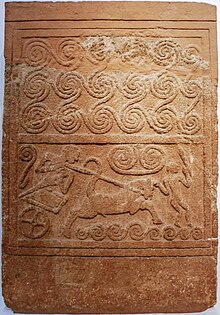 A limestone grave marker, decorated with spirals at the top and with a scene of a chariot-mounted warrior running down an enemy at the bottom