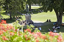 Studierende der Universität Bern sitzen auf der Grossen Schanze im Rasen unter Bäumen, in der Mitte das Hallerdenkmal, im Vordergrund Geranien
